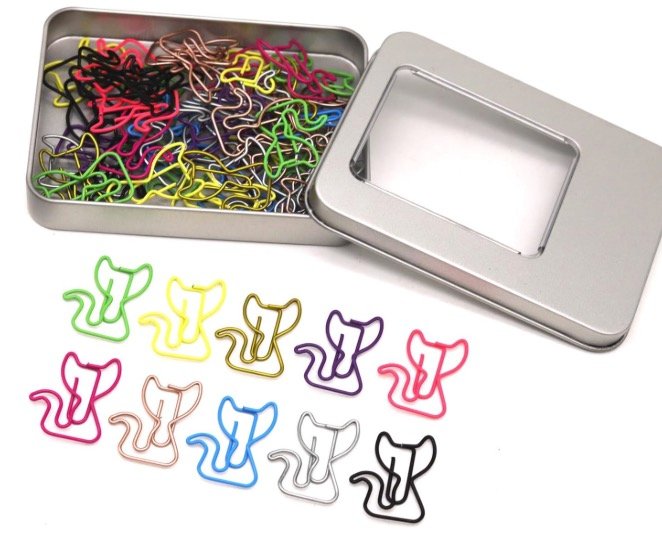 Colorful cat-shaped metal paper clips in a silver tin with a clear window lid, with assorted clips arranged in the foreground.